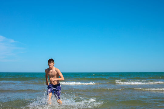 Shirtless teenage boy in swim shorts running out of sea waves toward camera, blue sky above, green water and horizon behind