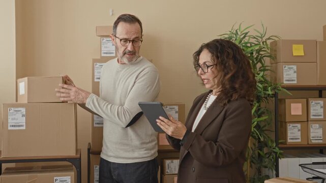 Man holds parcel and gestures with pen while woman checks tablet amid stacked boxes in a building; focused teamwork.