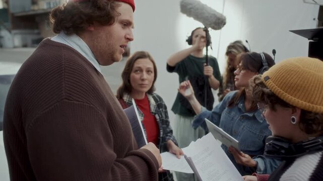 Behind the scenes of movie production crew preparing for outdoor shoot, female director discussing script with actors while makeup artist applying final touches and videographer positioning camera rig