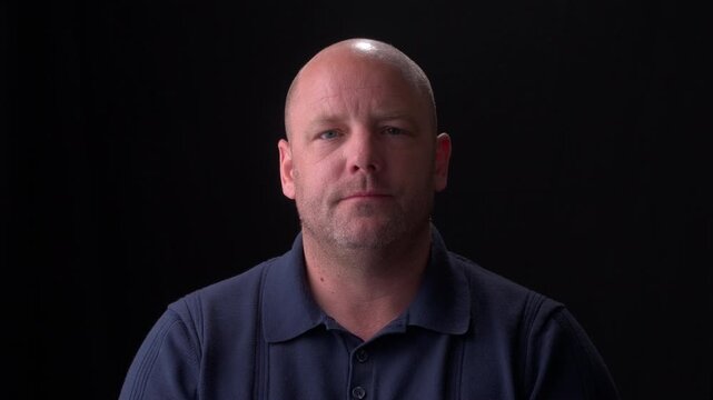 Middle-aged white bald man wearing a dark blue collared shirt with a plain black background, expressionless. 