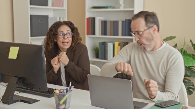Man and woman gesture with raised hands and playful fist pumps at a computer monitor and laptop in an office; celebration teamwork.