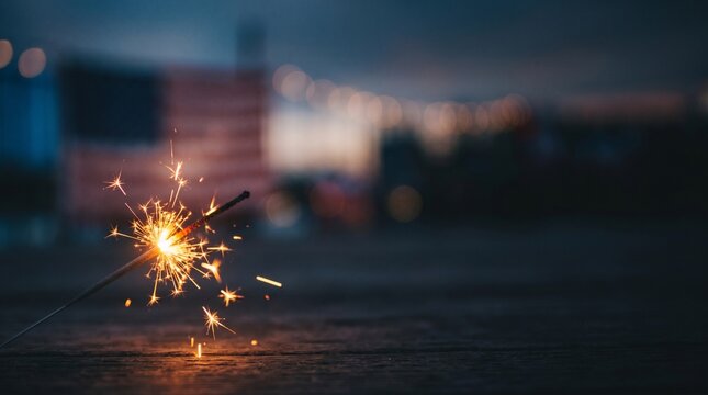 Sparkler burning at dusk with American flag and bokeh lights in background