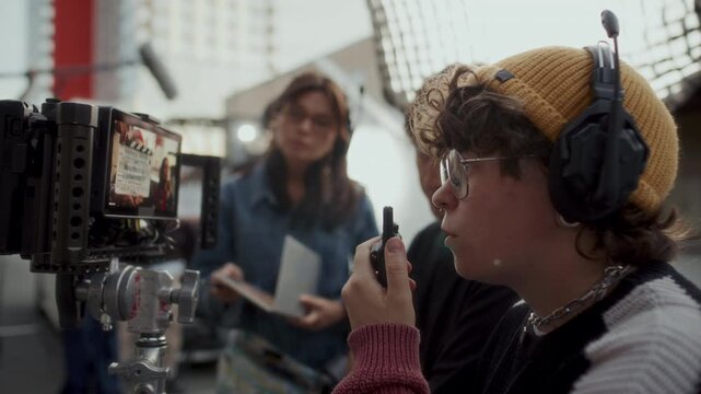 Young female film director in headset using walkie talkie to coordinate crew and watching camera monitor during shoot on outdoor movie set
