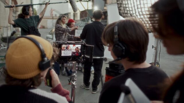 Film crew members wearing headsets monitoring actors, using walkie talkie and discussing scene on video camera screen during shoot on outdoor movie set. Over the shoulder view