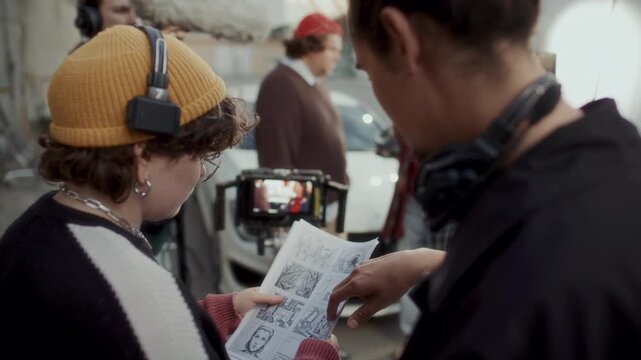 Behind the scenes shot of female movie director and cameraman discussing storyboard sketches during video shoot on outdoor set, boom operator and actors standing in background