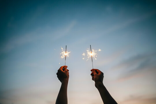 Hands holding two sparklers against twilight sky outdoors.
