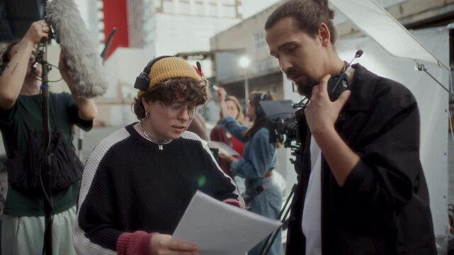 Young female film director and cameraman discussing script pages on outdoor movie set while boom operator, actors, stylist and clapper loader working in background