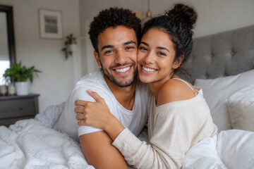 Obraz premium Two people are smiling and hugging each other while sitting in bed. The room has soft lighting that highlights their happy expressions. Plants are visible in the background.