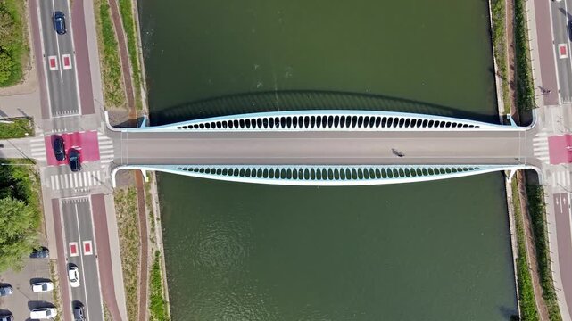 Aerial top-down drone view of a white arched bridge crossing a green canal with roads crosswalks and cars on both banks while rowers train on the water a summer view of urban infrastructure.