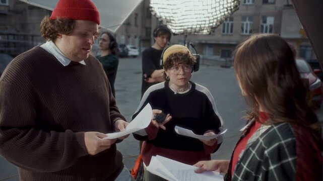 Female filmmaker in headset explaining script details and giving directions to actor and actress on outdoor movie set, cameraman and boom operator setting up equipment in background
