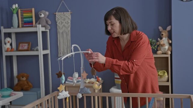 Woman adjusting a hanging mobile over a wooden cradle in a baby nursery with toys, shelves, and a macrame wall hanging; tenderness bonding.