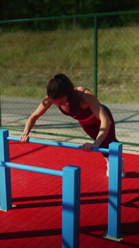A fit young woman performs push-ups on parallel bars on an outdoor sports ground. She engages in a focused and determined fitness training session.