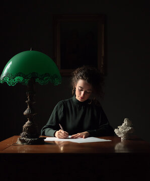 Imagen de una mujer concentrada escribiendo una carta sobre un escritorio de madera cl&aacute;sico, iluminada por luz &iacute;ntima de una antigua l&aacute;mpara verde de banquero en un ambiente oscuro. Romanticismo XIX