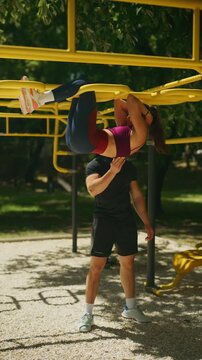 A woman performs an intense abdominal workout, hanging upside down with the support of a man. They are exercising together on monkey bars in a sunny outdoor gym.