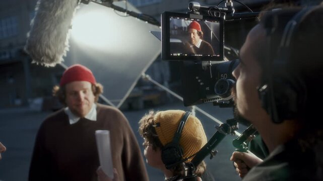 Camera operator reviewing composition on field monitor screen while female film director and actors discussing scene during outdoor shoot