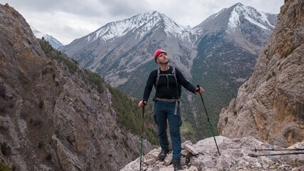 A person stands on a rocky peak looking up at the sky. The mountains rise in the background, showing snow on their tops. The scene shows a strong connection to nature during a hiking trip. © Evgeniy