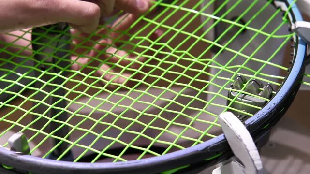 Tennis racket being restrung with bright green tennis strings on stringing machine, hands interlock overlapping main strings during installation. Regular maintenance of sports gear, weaving