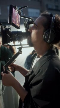Focused male camera operator in communication headset operating camera rig and watching external field monitor displaying live take, film crew working in background. Vertical side view