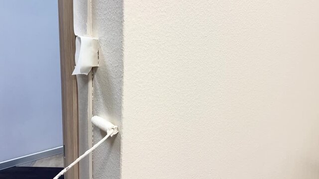 Worker's hand using paint roller, painting corner of wall near door frame with light paint during renovation in office or apartment, copy space. Handyman, repair job, maintenance