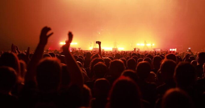 A large crowd of people standing tightly together and clapping their hands during a live performance lit by an intense orange glow.