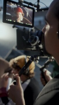 Professional camera operator reviewing actor performance on external monitor during video production shoot at outdoor location.Vertical behind the scenes shot