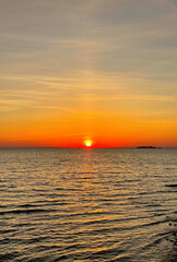 Obraz premium Sun dips below horizon over calm waters, casting warm orange glow across sky and sea. Serene sunset marks end of day.