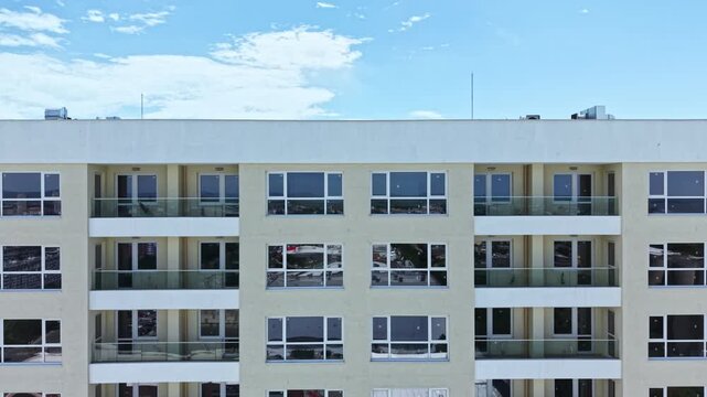 An apartment building stands with multiple levels. Each floor has many windows with reflections of clouds and the sky. Balconies extend out from the walls.