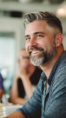 Obraz premium Confident middle aged man with stylish gray hair and beard smiling during a casual business meeting in a modern office. Natural light highlights his positive expression and charisma