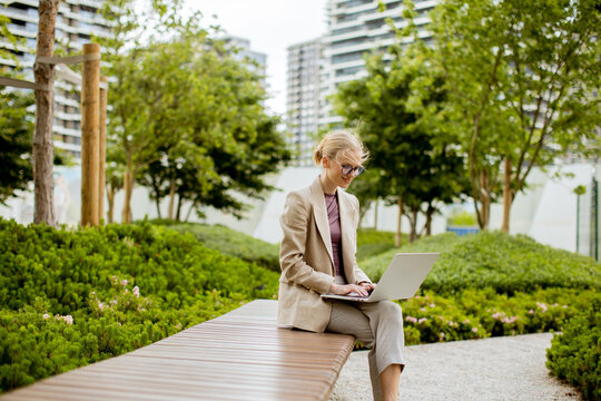 Woman working on laptop in urban park surrounded by greenery and buildings