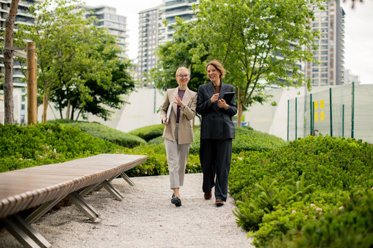 Two women walk through a park in a city while discussing work or projects