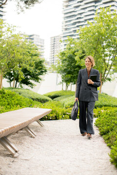 Woman walks through green space in city while carrying a black bag near buildings
