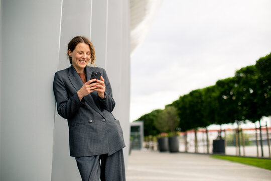 Woman in business suit smiles while texting on smartphone outside during day