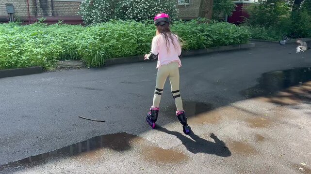a young girl from behind as she rollerblades along a paved path in a sunny, residential courtyard. She is wearing a bright pink helmet and full protective gear
