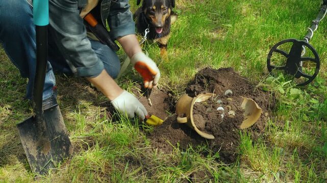 Person or archaeologist digging in the ground with a trowel, uncovering a ceramic pot and small objects while a dog watches nearby in a grassy outdoor setting