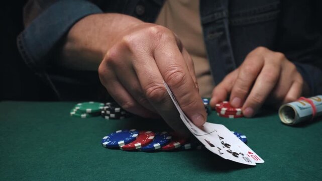 A male poker player with concentration shows a two pairs card combination, placing colorful chips on a green felt table in a dimly lit gaming room filled with foreboding
