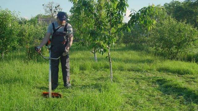 Man gardener in workwear trims grass with a string trimmer in a lush garden surrounded by trees and greenery under bright sunlight