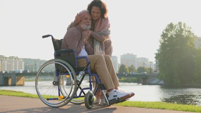 Old man in wheelchair walking with caregiver senior woman on road in park. Elderly family couple woman supporting embracing paralyzed man in chair for people with disability outdoor. Rehabilitation