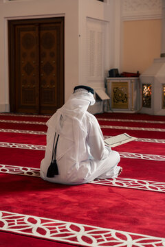 Person in white traditional clothing reading a book on a red patterned carpet.