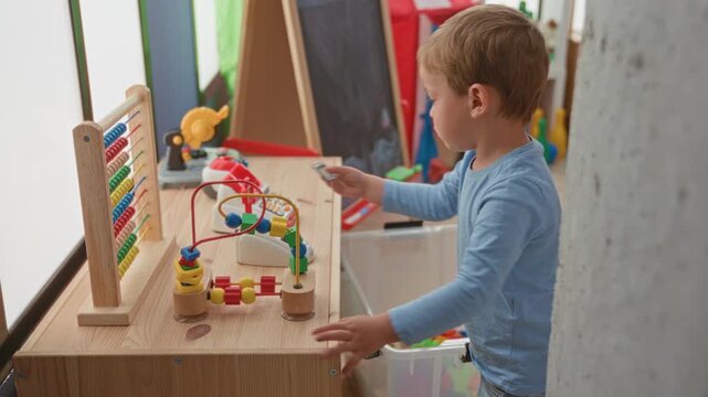 Boy reaching hand to wooden bead maze and abacus on a table in a playroom, toy bin and chalkboard nearby; curiosity learning play.