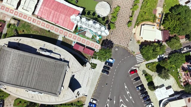 Bird's eye view shows a busy city scene with cars parked near a large building surrounded by trees. Streets connect different areas, and paths are lined with greenery.