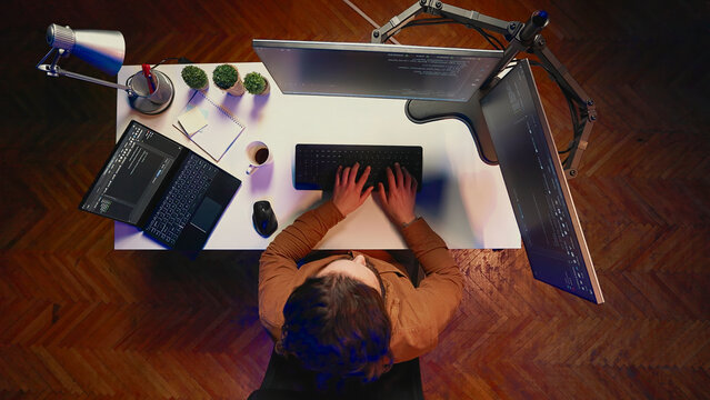 Top-down view of remote worker typing code in personal office, using JavaScript language to develop software application. IT specialist using multi-monitor desktop setup to finish project.