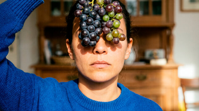 Pensive woman holds clustered grape over face in intimate home portrait
