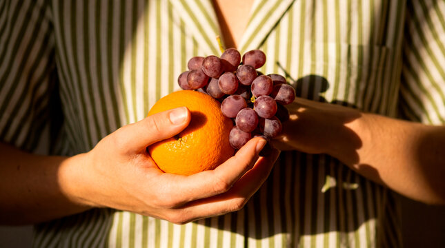 Attentive hands cradle orange and grape in striped shirt under warm sunlight