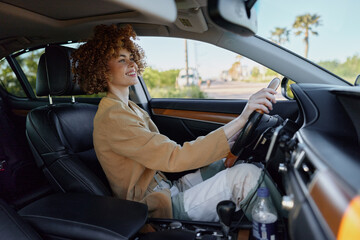 Naklejka premium Smiling woman with curly hair driving a car, wearing a beige jacket. Close up on interior with natural light. Concept of lifestyle, travel, and self expression. Retro film color adds warmth.