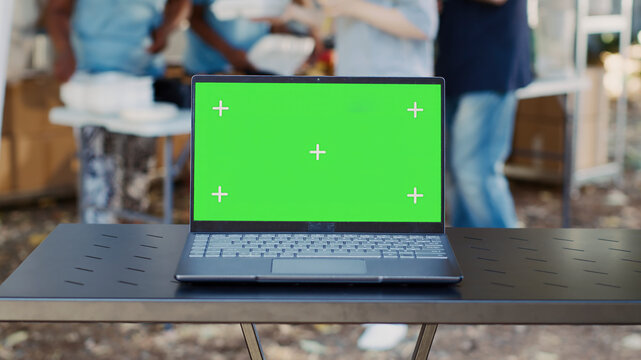 Electronic laptop with isolated green screen sits on a table while charity workers assist the less privileged, detailed view of minicomputer with a blank copyspace mockup template.