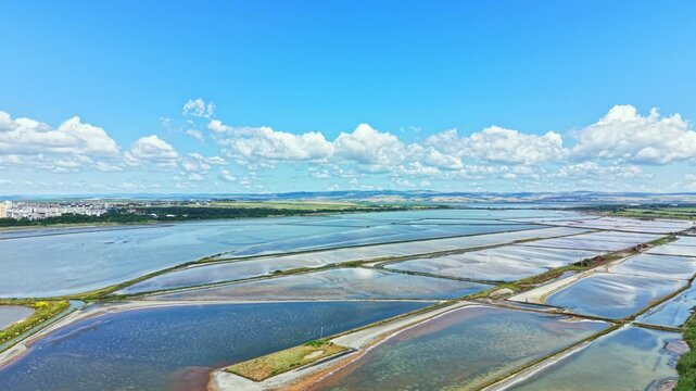 The salt pans in Burgas are visible with water reflecting the sky. The landscape stretches out with rows of salt ponds and distant hills under a sunny sky full of clouds during summer.
