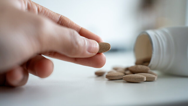 Hand picking single brown supplement tablet from white surface near open bottle
