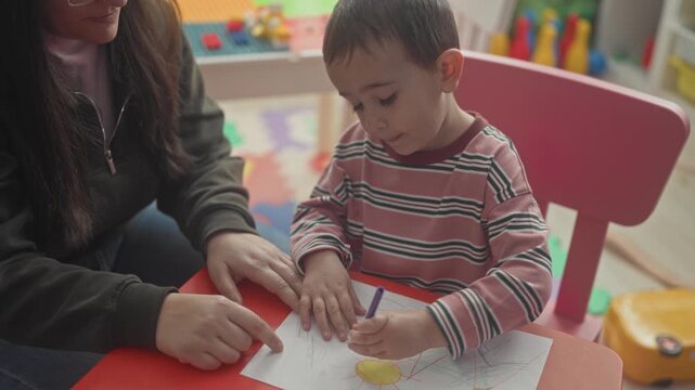 Woman teacher guides toddler's hand holding a purple crayon while the little boy draws scribbles on paper at a preschool playroom table; learning concentration.