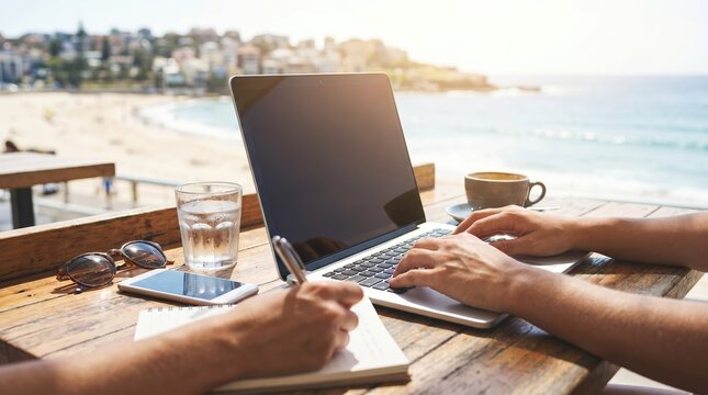 Digital nomad working on laptop at beach cafe, young man typing on computer and writing in notebook, remote work lifestyle concept