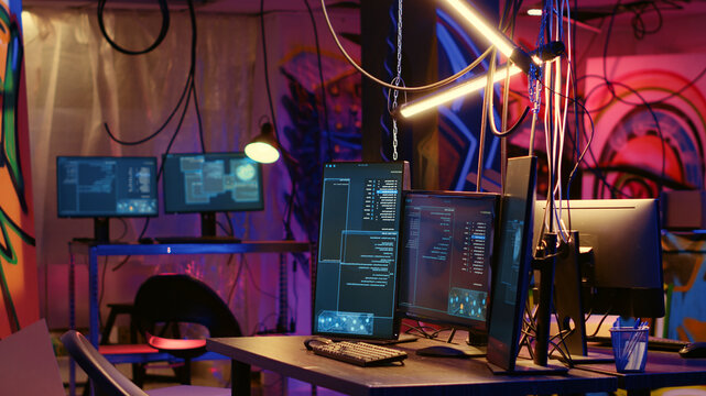 Programming language on PC screens in empty messy hackers base of operations with grunge graffiti drawings sprayed on walls, Neon lit ghetto hideout used by criminals to commit illegal activities.